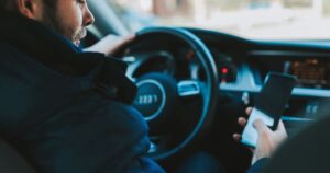 A man in a coat, sitting in the driver's seat of a vehicle looking at a phone in his hand