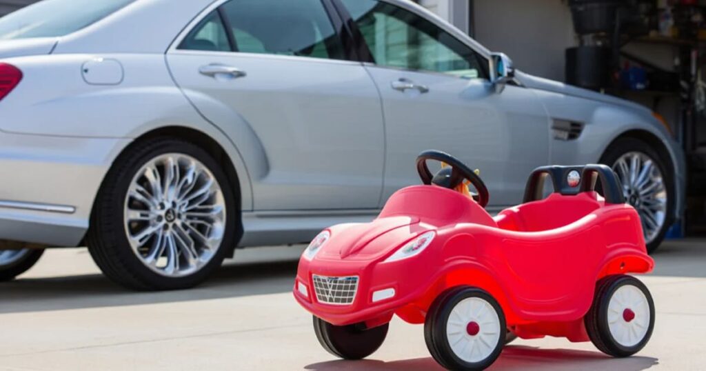 A small red kids car on a driveway next to a white vehicle
