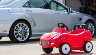 A small red kids car on a driveway next to a white vehicle